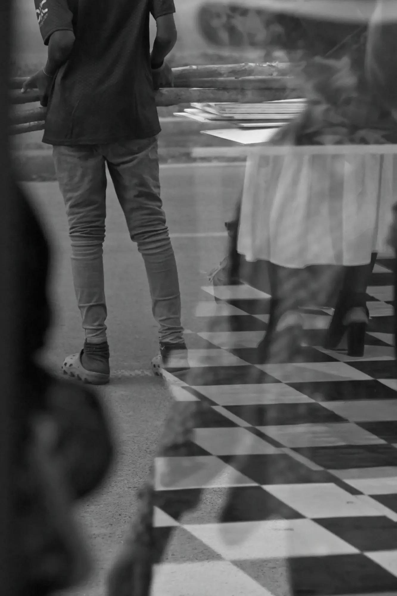 A figure in jeans at a timber rail, seen through café glass with a chequered floor and passer-by reflected