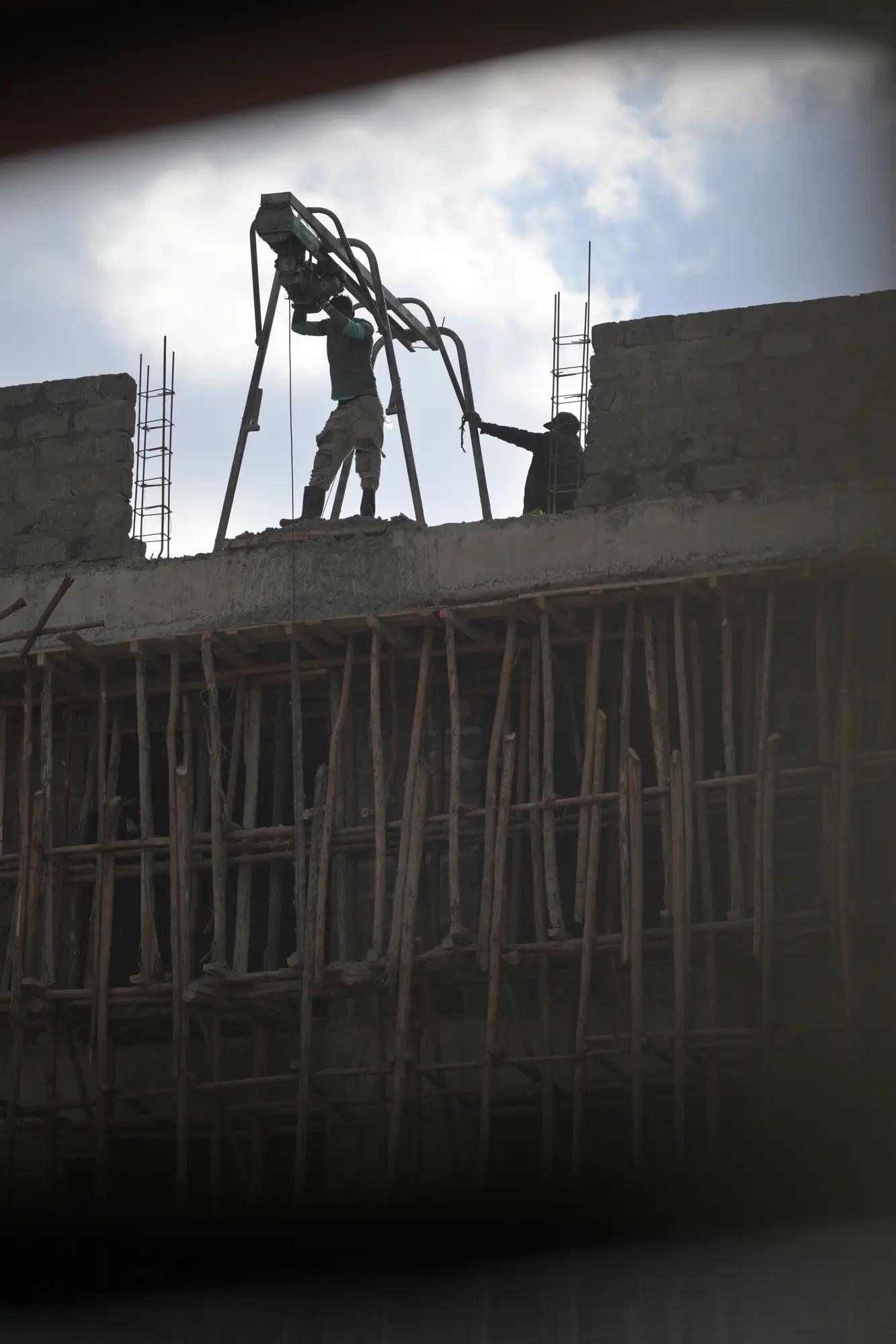 Two builders silhouetted on an unfinished slab above a dense forest of timber scaffolding poles