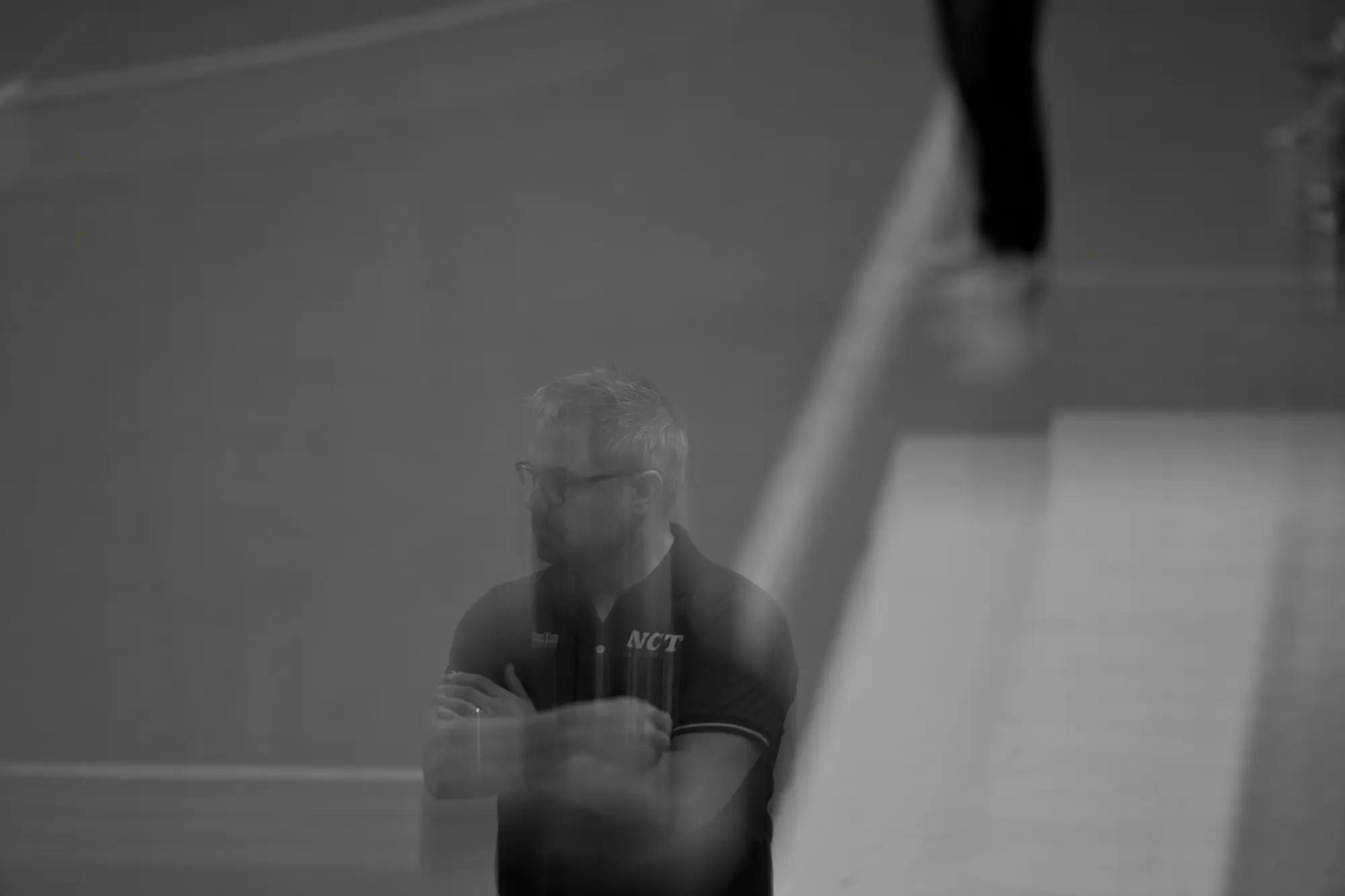 A coach stands arms folded and ghosted by movement, alone in a wide empty stretch of court